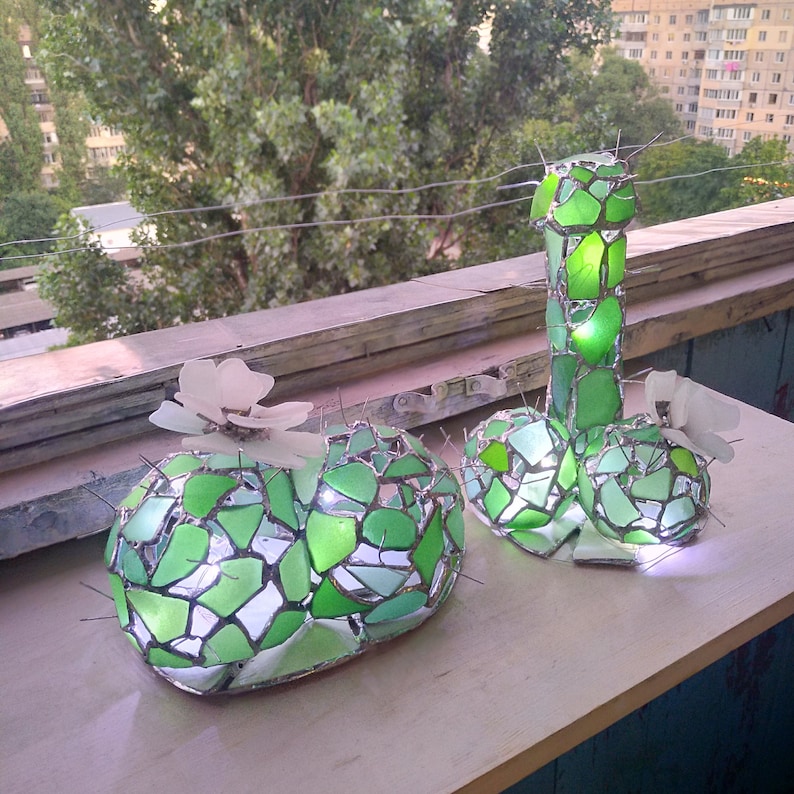 May include: Two green glass mosaic cactus sculptures with white flowers. The cactus on the right is tall and slender, while the cactus on the left is round and flat. Both sculptures are illuminated from within.