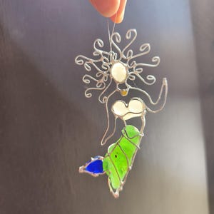 Puede incluir: Adorno de sirena de vidriera hecho a mano. La sirena tiene un marco de alambre plateado con cabeza y pecho blancos, cuerpo verde y cola azul. El adorno se sujeta con un alambre fino.