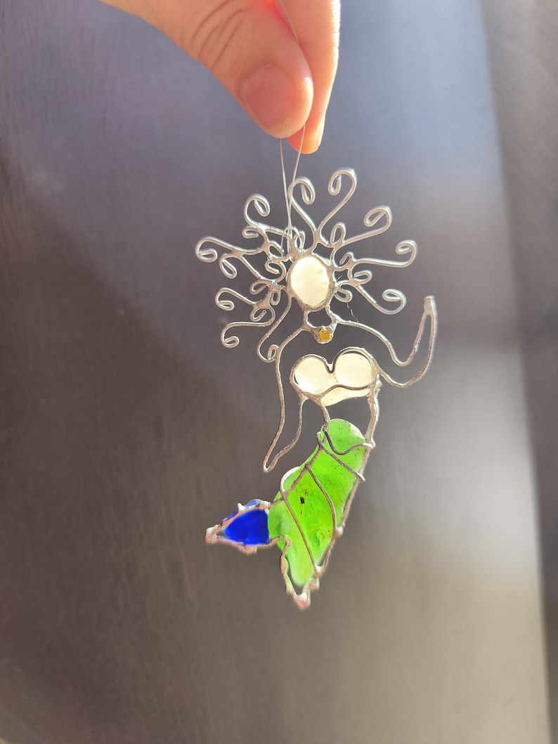 Puede incluir: Adorno de sirena de vidriera hecho a mano. La sirena tiene un marco de alambre plateado con cabeza y pecho blancos, cuerpo verde y cola azul. El adorno se sujeta con un alambre fino.