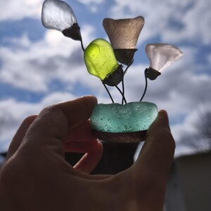 May include: A handmade sculpture of sea glass flowers. The flowers are crafted from different colours of sea glass, including clear, green, and pink. The flowers are attached to thin, dark stems and set in a turquoise sea glass base.