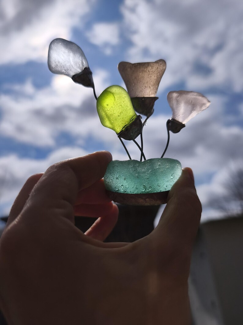 May include: A handmade sculpture of sea glass flowers. The flowers are crafted from different colours of sea glass, including clear, green, and pink. The flowers are attached to thin, dark stems and set in a turquoise sea glass base.