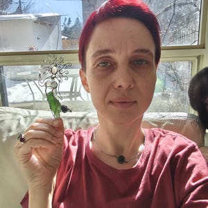 Puede incluir: Una escultura de sirena de alambre con cola verde y detalles blancos se sostiene frente a una ventana. La sirena tiene un dise&ntilde;o fluido y est&aacute; hecha de metal. El fondo incluye una ventana, un sof&aacute; y una persona con una camiseta roja.