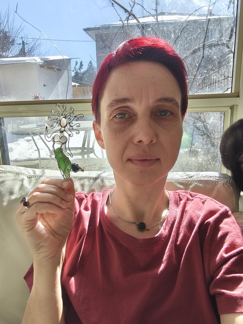 Puede incluir: Una escultura de sirena de alambre con cola verde y detalles blancos se sostiene frente a una ventana. La sirena tiene un dise&ntilde;o fluido y est&aacute; hecha de metal. El fondo incluye una ventana, un sof&aacute; y una persona con una camiseta roja.