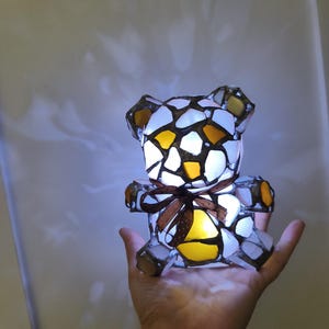 May include: A stained glass teddy bear lamp with a brown ribbon around its middle. The bear is made of white, yellow, and brown glass pieces. The lamp is lit from within, creating a warm glow.