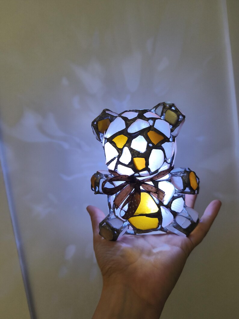 May include: A stained glass teddy bear lamp with a brown ribbon around its middle. The bear is made of white, yellow, and brown glass pieces. The lamp is lit from within, creating a warm glow.