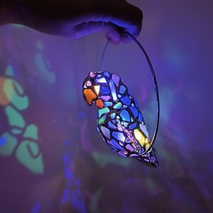 May include: A colorful, mosaic-style parrot-shaped lamp with a metal hanging loop. The lamp features a vibrant array of colors, including blue, purple, orange, and yellow. The light shines through the mosaic pieces, creating a beautiful display of light and shadow.