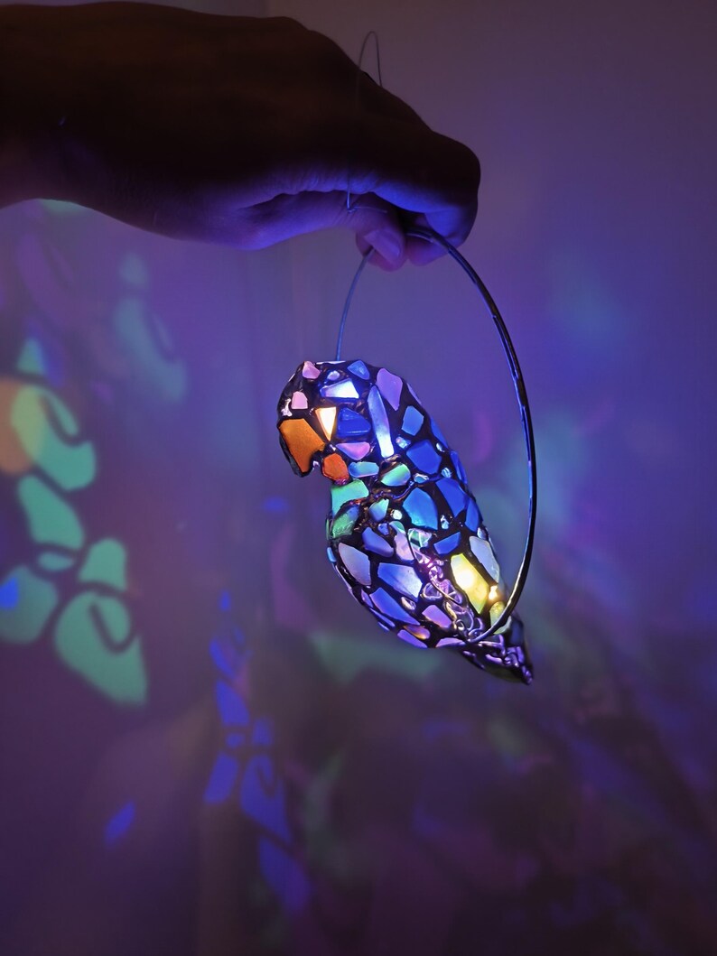 May include: A colorful, mosaic-style parrot-shaped lamp with a metal hanging loop. The lamp features a vibrant array of colors, including blue, purple, orange, and yellow. The light shines through the mosaic pieces, creating a beautiful display of light and shadow.
