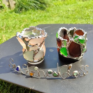 May include: Two small glass candle holders made with sea glass. The candle holders are on a black surface with the words "Sea Glass" written in wire with sea glass accents.