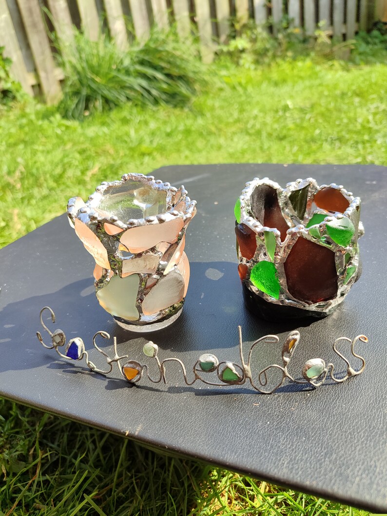 May include: Two small glass candle holders made with sea glass. The candle holders are on a black surface with the words "Sea Glass" written in wire with sea glass accents.