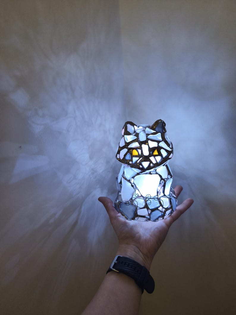 May include: A cat-shaped mosaic lamp emitting a warm light. The lamp is made from irregular pieces of white, blue, and brown glass, giving it a unique texture. Light shines through the glass, illuminating the room. The lamp is held in a hand.