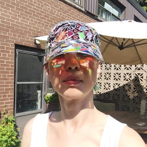 May include: A colorful stained glass hat with a brim. The hat's design includes red, orange, yellow, green, blue, and purple glass pieces. The sunlight casts colorful shadows on the person's face, creating a vibrant effect.