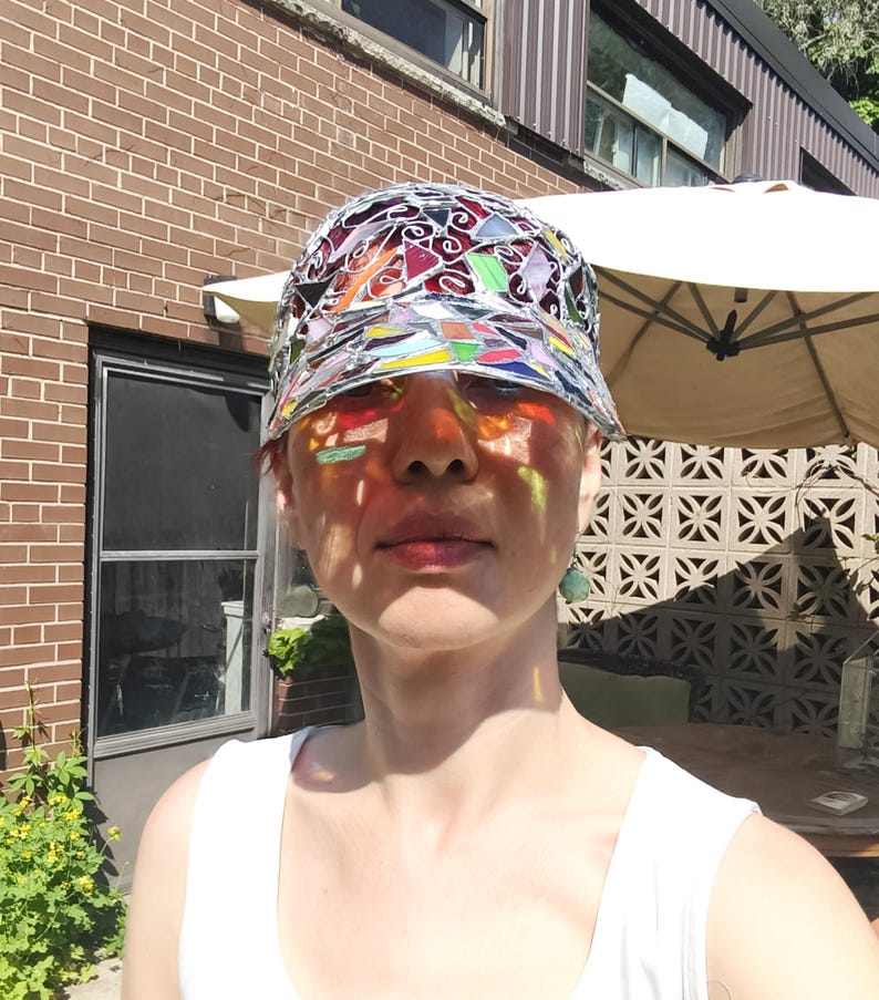 May include: A colorful stained glass hat with a brim. The hat's design includes red, orange, yellow, green, blue, and purple glass pieces. The sunlight casts colorful shadows on the person's face, creating a vibrant effect.