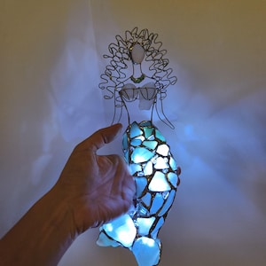 Puede incluir: Una escultura decorativa de sirena hecha de alambre y vidrio marino. La figura presenta una cabeza y un torso formados por alambre con detalles de vidrio, y una cola construida con m&uacute;ltiples piezas de vidrio azul iluminadas por luces internas.