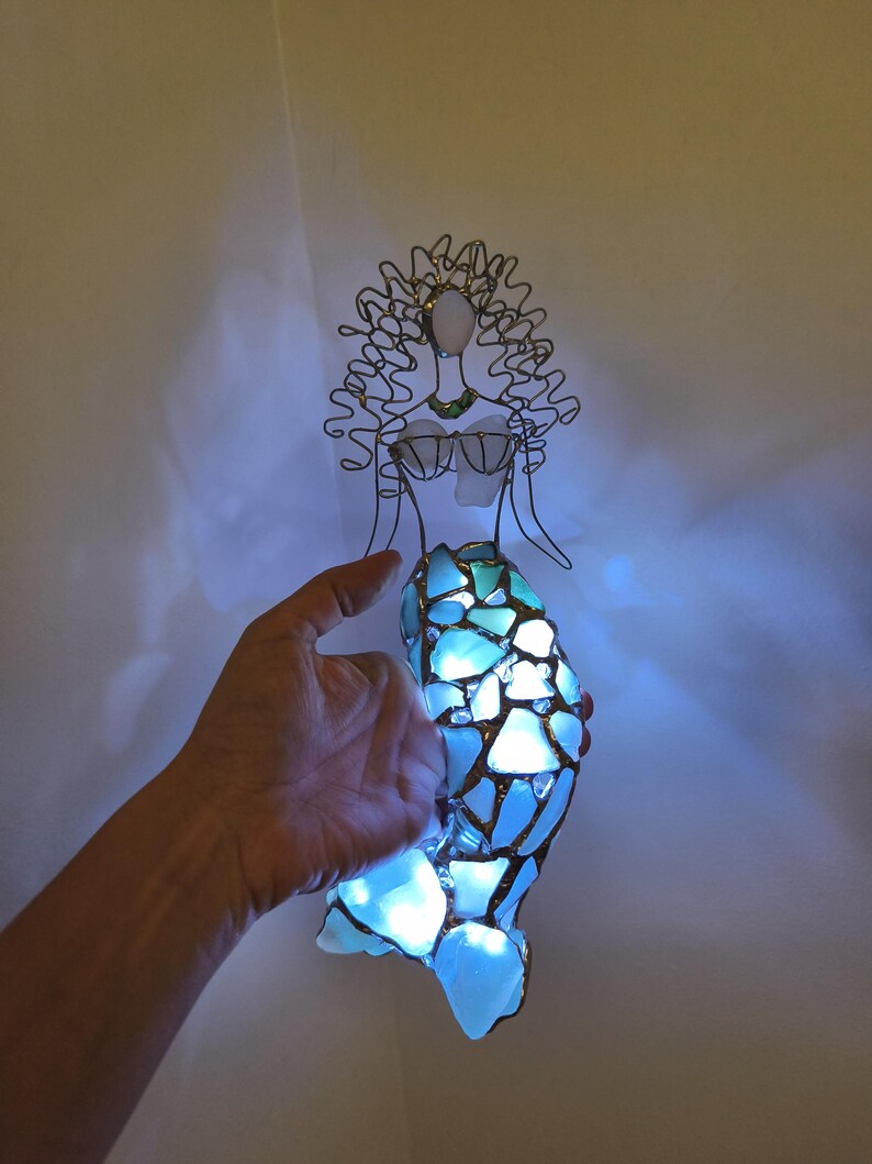 Puede incluir: Una escultura decorativa de sirena hecha de alambre y vidrio marino. La figura presenta una cabeza y un torso formados por alambre con detalles de vidrio, y una cola construida con m&uacute;ltiples piezas de vidrio azul iluminadas por luces internas.