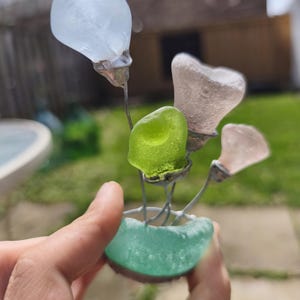 May include: A handcrafted sea glass flower sculpture. The sculpture features a teal base holding wire stems with various sea glass flower shapes in white, pink, and green. The sculpture is held in a hand.