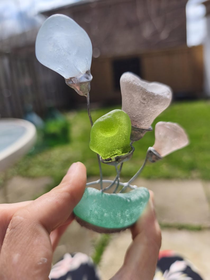 May include: A handcrafted sea glass flower sculpture. The sculpture features a teal base holding wire stems with various sea glass flower shapes in white, pink, and green. The sculpture is held in a hand.