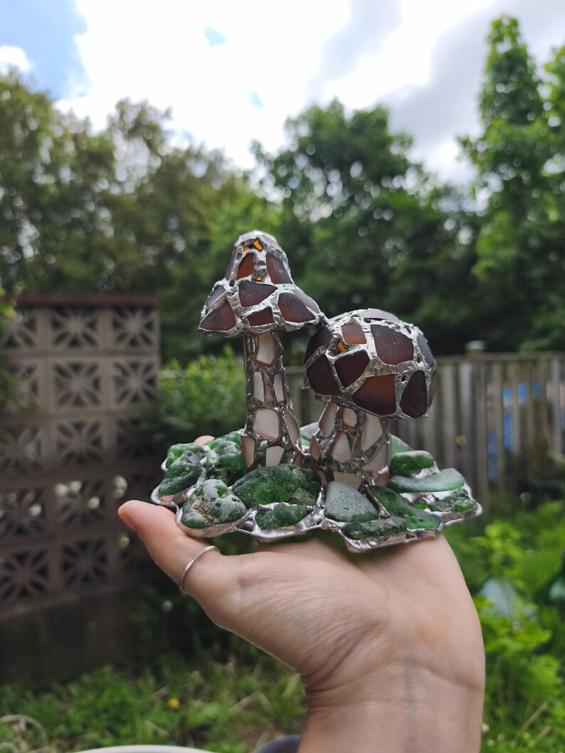May include: A handcrafted glass mushroom sculpture with brown and clear mosaic caps, white stems, and a base of green glass pieces. The sculpture is held in a hand against a blurred background of greenery.