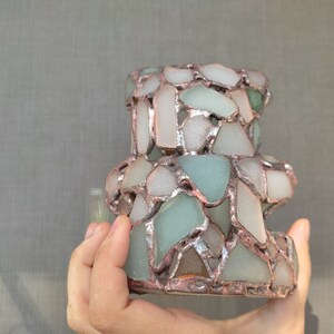 May include: A handcrafted, decorative candle holder made from sea glass pieces in shades of white, green, and blue. The sea glass is held together with a copper-colored metal. The candle holder has a unique, textured appearance.