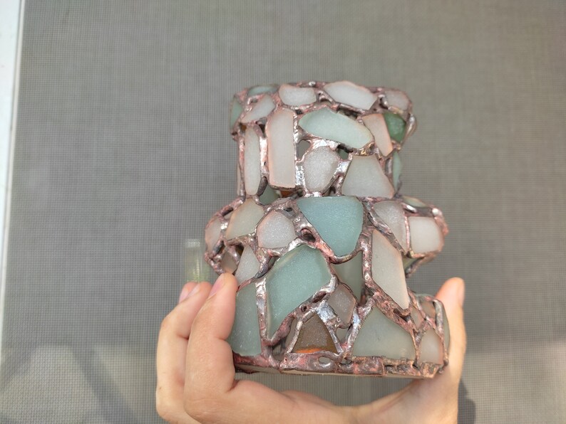 May include: A handcrafted, decorative candle holder made from sea glass pieces in shades of white, green, and blue. The sea glass is held together with a copper-colored metal. The candle holder has a unique, textured appearance.