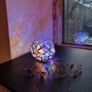 May include: A stained glass lamp with a colorful light cast on a black surface. The word "Seastone" is spelled out in wire with colorful stones.