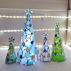 Pode incluir: Uma cole&ccedil;&atilde;o de cinco &aacute;rvores de Natal decorativas. Cada &aacute;rvore, em forma de cone, &eacute; feita de vidro do mar em tons de azul, verde e branco. Cada &aacute;rvore &eacute; encimada por uma estrela e iluminada com luzes internas. As &aacute;rvores variam em tamanho.