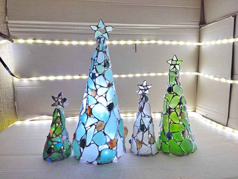 Pode incluir: Uma cole&ccedil;&atilde;o de cinco &aacute;rvores de Natal decorativas. Cada &aacute;rvore, em forma de cone, &eacute; feita de vidro do mar em tons de azul, verde e branco. Cada &aacute;rvore &eacute; encimada por uma estrela e iluminada com luzes internas. As &aacute;rvores variam em tamanho.