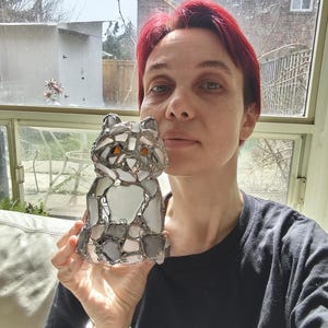 May include: A mosaic cat ornament, constructed from grey, white, and clear glass pieces. The cat has orange eyes. The decorative figurine is held up in front of a window. The cat is a decorative item.