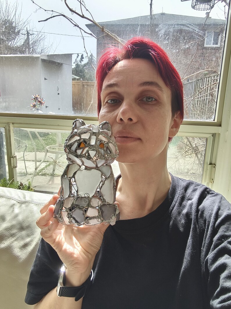 May include: A mosaic cat ornament, constructed from grey, white, and clear glass pieces. The cat has orange eyes. The decorative figurine is held up in front of a window. The cat is a decorative item.