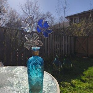 May include: A turquoise glass vase with a textured surface holds three decorative glass flowers. One flower is blue, another is clear, and the third is a butterfly shape. The vase sits on a glass table outdoors.