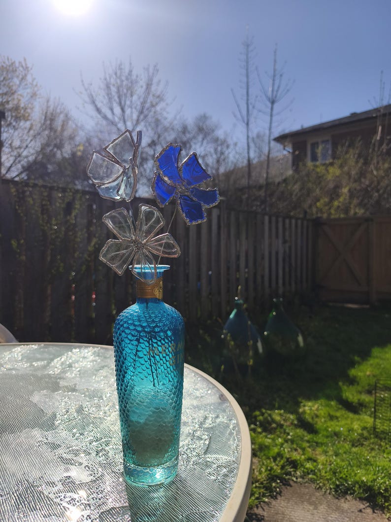 May include: A turquoise glass vase with a textured surface holds three decorative glass flowers. One flower is blue, another is clear, and the third is a butterfly shape. The vase sits on a glass table outdoors.