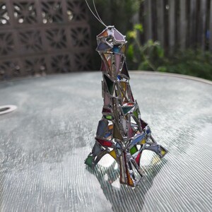 May include: A stained glass Eiffel Tower ornament in a variety of colours. The miniature tower is constructed from geometric shapes of coloured glass, including pink, blue, green, and yellow, joined with silver solder. A thin wire is attached for hanging.
