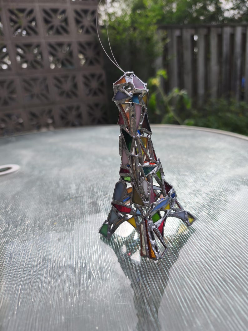 May include: A stained glass Eiffel Tower ornament in a variety of colours. The miniature tower is constructed from geometric shapes of coloured glass, including pink, blue, green, and yellow, joined with silver solder. A thin wire is attached for hanging.