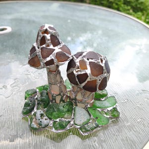 May include: A handcrafted glass art piece featuring two mushroom sculptures. The mushroom caps are made of brown glass pieces, and the stems are silver. The base is composed of green glass pieces, creating a whimsical, nature-inspired design.