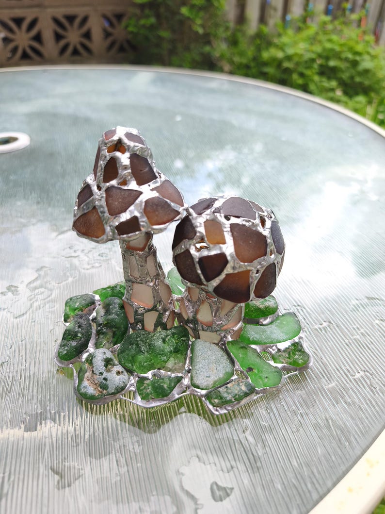 May include: A handcrafted glass art piece featuring two mushroom sculptures. The mushroom caps are made of brown glass pieces, and the stems are silver. The base is composed of green glass pieces, creating a whimsical, nature-inspired design.