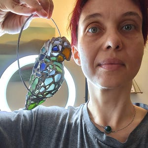 May include: A stained glass parrot sculpture with a wire frame. The bird features a mosaic of blue, green, and amber glass pieces. The parrot is suspended from a wire loop. The background includes a ring light and a person.