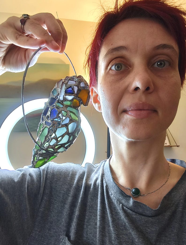 May include: A stained glass parrot sculpture with a wire frame. The bird features a mosaic of blue, green, and amber glass pieces. The parrot is suspended from a wire loop. The background includes a ring light and a person.