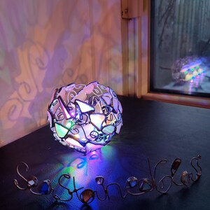 May include: A heart-shaped stained glass lamp with colorful glass pieces, casting a rainbow-colored light on the word "Sea Stones" made of wire and glass.
