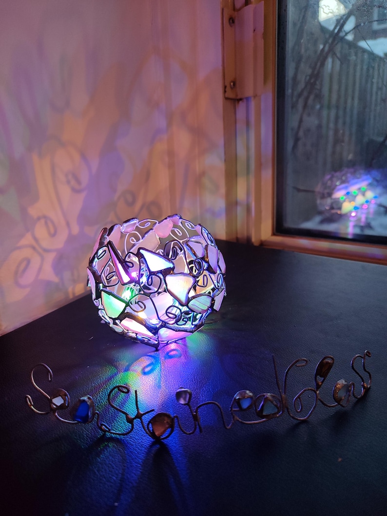 May include: A heart-shaped stained glass lamp with colorful glass pieces, casting a rainbow-colored light on the word "Sea Stones" made of wire and glass.