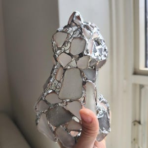 May include: A handcrafted cat sculpture made from sea glass pieces in white, grey, and clear hues. The sea glass is held together with silver-coloured metal. The cat sculpture is held in a person's hand.
