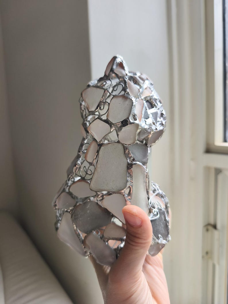 May include: A handcrafted cat sculpture made from sea glass pieces in white, grey, and clear hues. The sea glass is held together with silver-coloured metal. The cat sculpture is held in a person's hand.