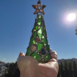 Pode incluir: Um enfeite de &aacute;rvore de Natal artesanal feito de peda&ccedil;os de vidro verde, conchas e um topo em forma de estrela. A &aacute;rvore &eacute; exibida contra um c&eacute;u azul brilhante, sugerindo um tema costeiro ou de praia. O enfeite tem cerca de 15 cm de altura.