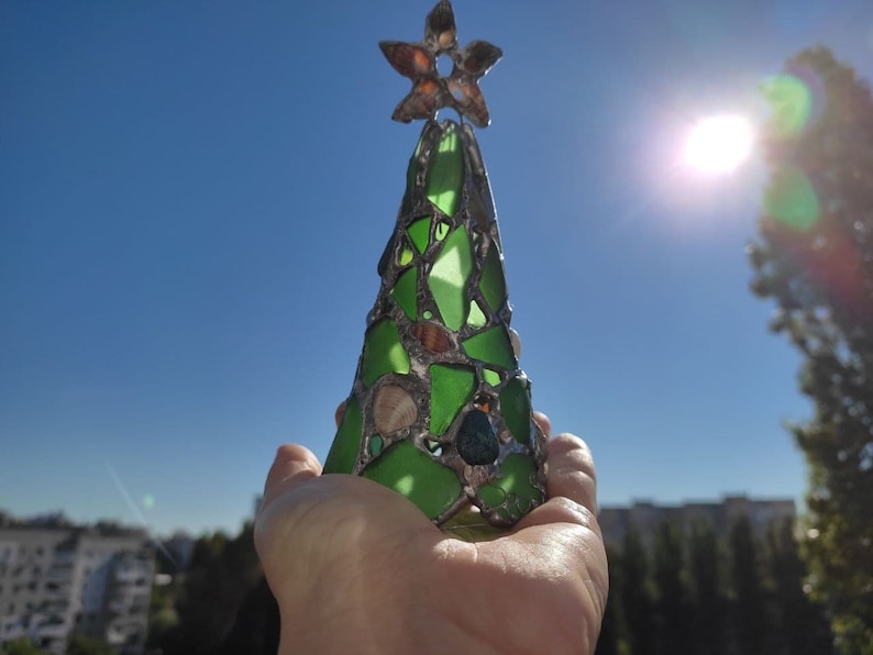 Pode incluir: Um enfeite de &aacute;rvore de Natal artesanal feito de peda&ccedil;os de vidro verde, conchas e um topo em forma de estrela. A &aacute;rvore &eacute; exibida contra um c&eacute;u azul brilhante, sugerindo um tema costeiro ou de praia. O enfeite tem cerca de 15 cm de altura.