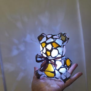 May include: A mosaic-style teddy bear lamp with a brown ribbon around its neck. The lamp is made of white, yellow, and brown glass pieces. The lamp is lit, and the light shines through the glass. The lamp is held in a person's hand.