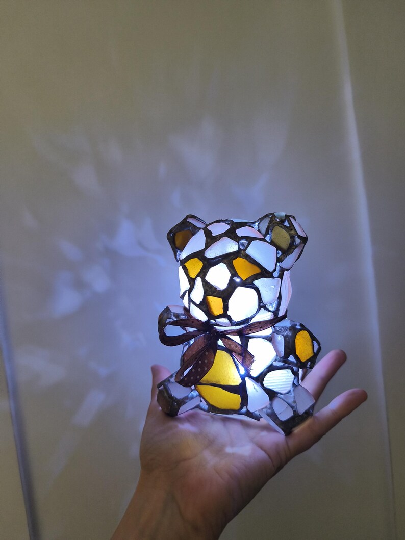 May include: A mosaic-style teddy bear lamp with a brown ribbon around its neck. The lamp is made of white, yellow, and brown glass pieces. The lamp is lit, and the light shines through the glass. The lamp is held in a person's hand.