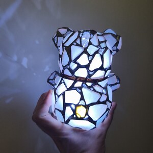 May include: A mosaic-style bear-shaped lamp with a warm, glowing light. The lamp is constructed from white and black pieces, creating a stained-glass effect. A brown band encircles the bear's midsection. The lamp is held in a hand.
