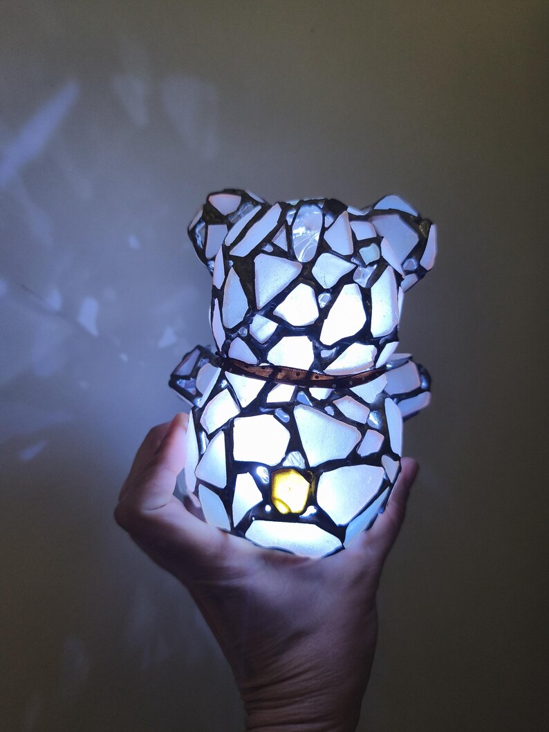 May include: A mosaic-style bear-shaped lamp with a warm, glowing light. The lamp is constructed from white and black pieces, creating a stained-glass effect. A brown band encircles the bear's midsection. The lamp is held in a hand.