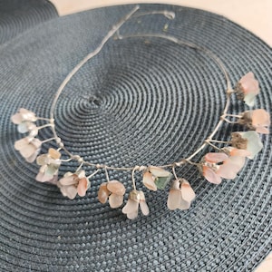 May include: A delicate silver-toned wire headband adorned with clusters of small, translucent, pink and green gemstone flowers. The floral accents are arranged along the wire, creating a subtle, elegant design. The headband rests on a textured, blue-gray surface.