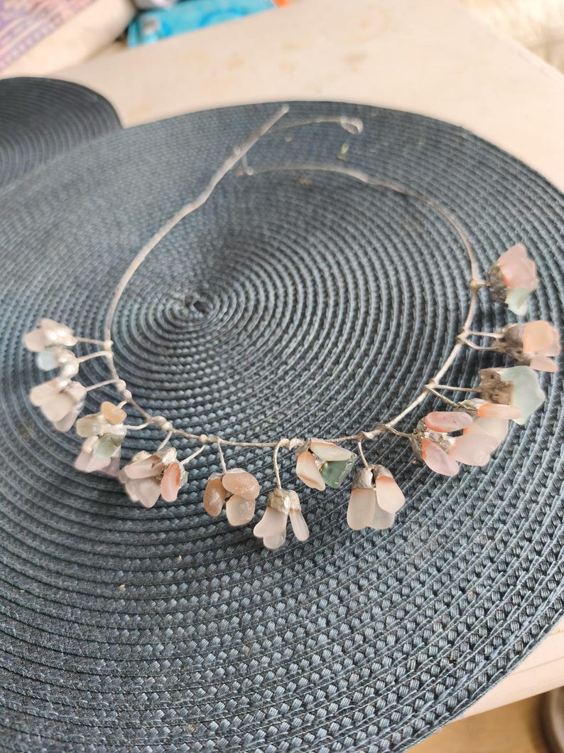 May include: A delicate silver-toned wire headband adorned with clusters of small, translucent, pink and green gemstone flowers. The floral accents are arranged along the wire, creating a subtle, elegant design. The headband rests on a textured, blue-gray surface.