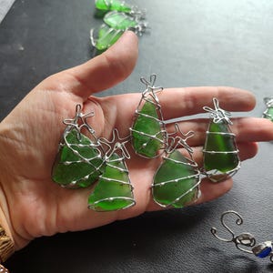 May include: Five green glass Christmas tree ornaments wrapped in silver wire. The ornaments are arranged in a hand.