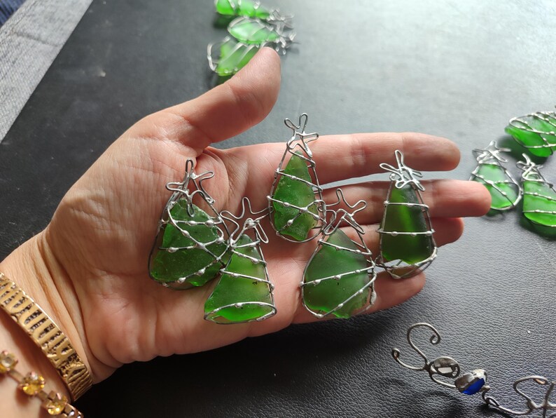 May include: Five green glass Christmas tree ornaments wrapped in silver wire. The ornaments are arranged in a hand.